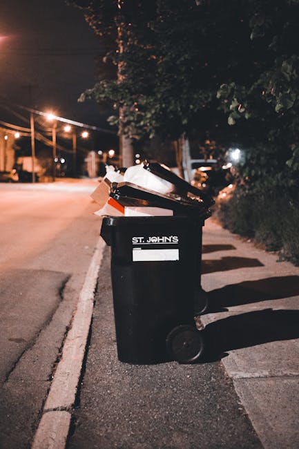 A black wheeled rubbish bin with a white label and the name 'St. John’s' printed on the front, standing on a dark asphalt sidewalk beside a curb. The bin's lid is open, revealing a mixture of discarde