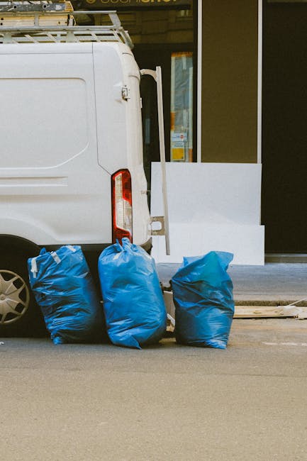 The image shows the rear section of a white commercial van parked on a paved street. Three large blue rubbish bags, made of thick plastic with a slightly wrinkled texture, are positioned on the ground