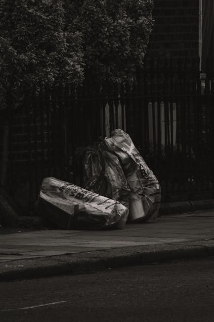 The image shows the rear section of a white commercial van parked on a paved street. Three large blue rubbish bags, made of thick plastic with a slightly wrinkled texture, are positioned on the ground