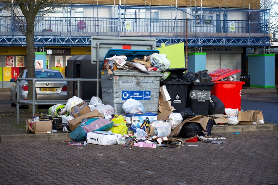 A collection of black rubbish bags and discarded packaging materials piled against a weathered red metal door in an urban setting. The bags are piled haphazardly, with some partially open revealing co
