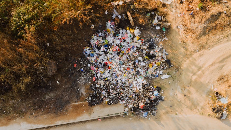 A large pile of mixed construction and household waste is visible in an outdoor setting, consisting of broken wood, scrap metal, plastic debris, cardboard, and other refuse. The materials are piled hi